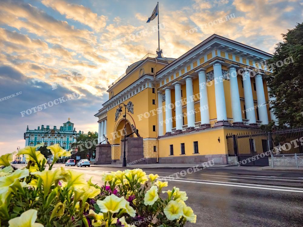 На фотографии изображены Адмиралтейство и Зимний дворец в Санкт-Петербурге. Величественные колонны Адмиралтейства освещены теплым светом заката, а на заднем плане виднеется зеленое здание Зимнего дворца. Летний вечер добавляет мягкий свет и романтическую атмосферу историческому центру города.
